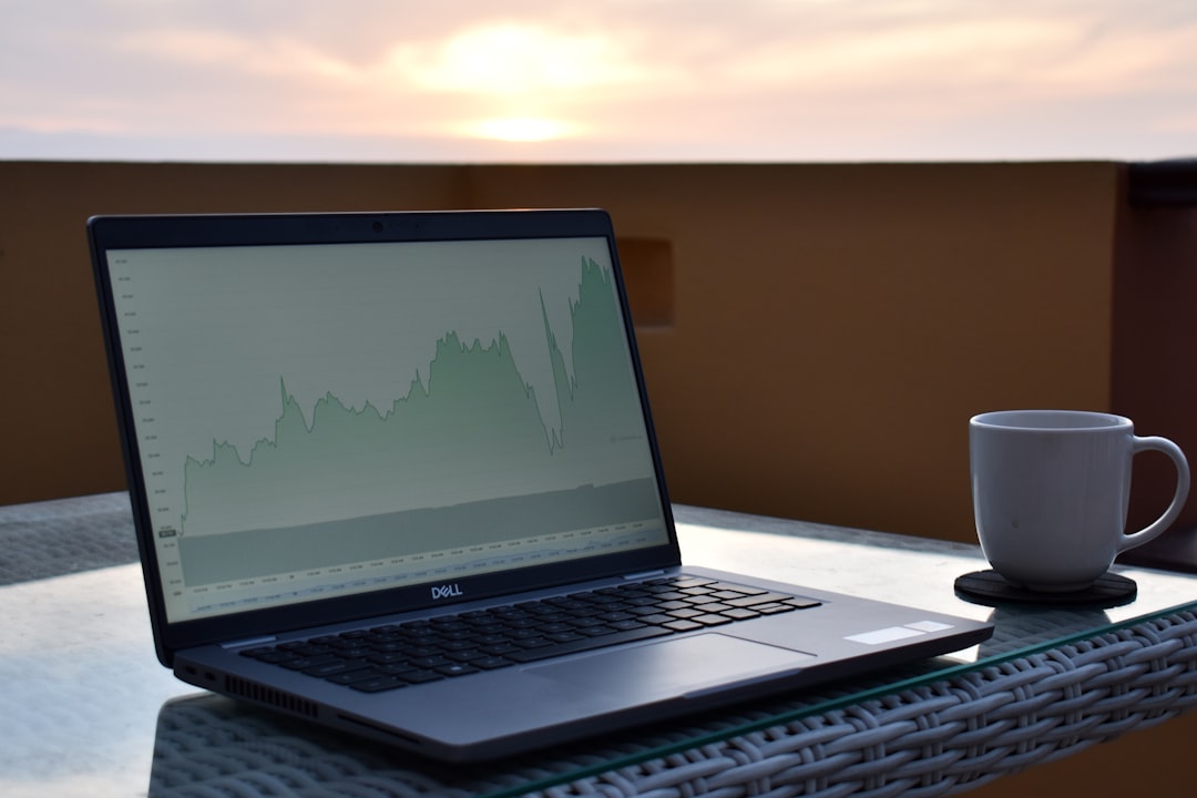 A laptop displaying a financial chart on a table with a coffee cup beside it against a sunset backdrop.